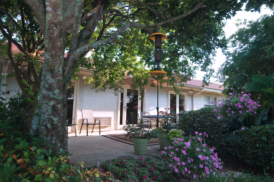 Outdoor patio area at Twin Oaks Nursing & Convalescent Home featuring a large tree, potted plants, a bird feeder, patio chairs, and a table with chairs surrounded by lush greenery and flowering plants.