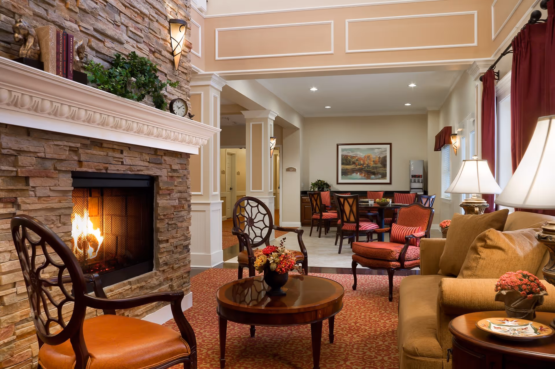 A cozy living room area in a senior living facility featuring a stone fireplace with a fire burning, two wooden chairs with orange cushions, a round wooden coffee table with a floral arrangement, a beige sofa with cushions, and a side table with a lamp and decorative items. In the background, there is a dining area with a table and chairs, a framed landscape painting on the wall, and a water dispenser.