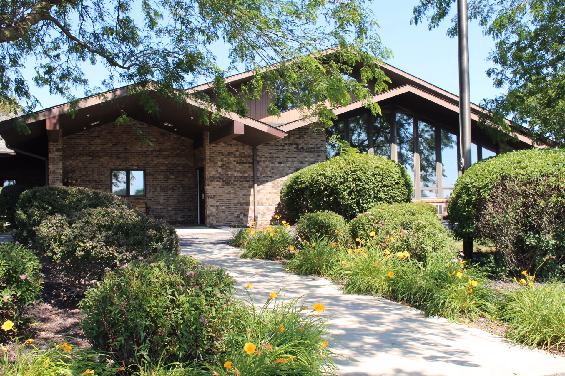Front entrance of a single-story brick building with a walkway, landscaped bushes and yellow flowers.