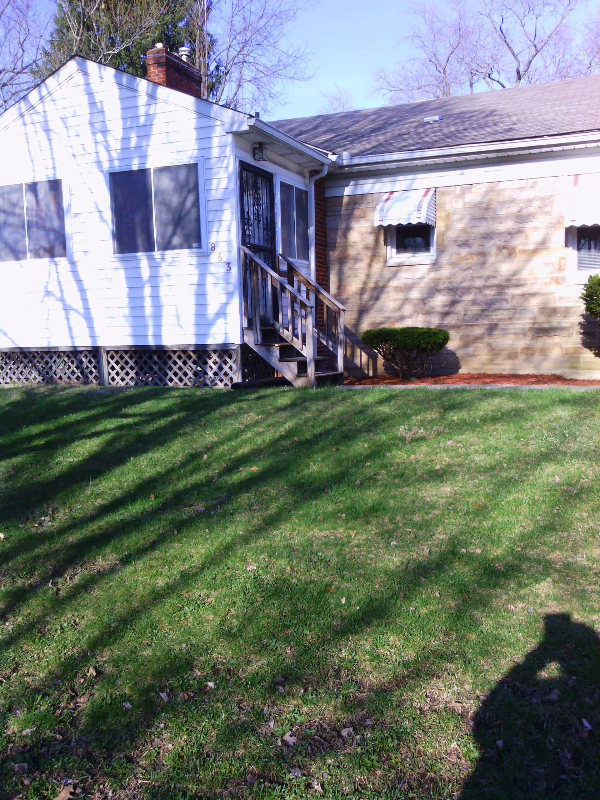 Exterior view of a single-story building with white siding and a stone facade. There is a small wooden staircase leading to a door with a metal security screen. The building has two windows with striped awnings and some small bushes near the foundation. The lawn in front is green with shadows of tree branches and a person taking the photo.