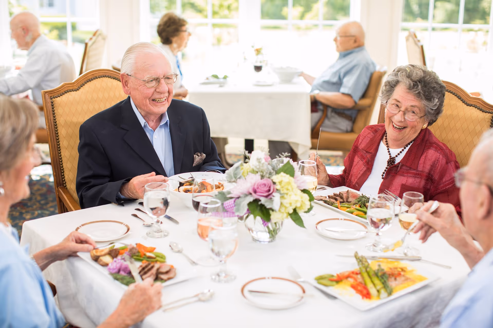 A group of elderly residents smiling and enjoying a meal together at a well-lit dining table with a floral centerpiece.