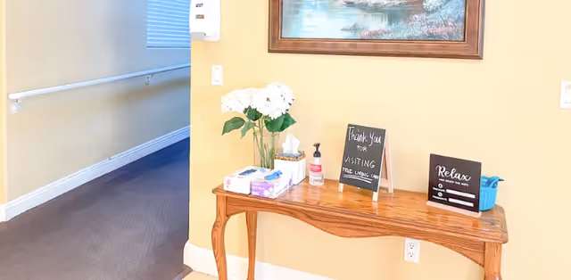 A hallway corner in a senior living facility with a wooden table against a yellow wall. On the table are a vase with white flowers, a box of tissues, hand sanitizer, and two small signs. One sign says 'Thank You for Visiting True Living Care' and the other says 'Relax and Enjoy the Day'. A framed painting hangs above the table, and a handrail is visible along the hallway wall.