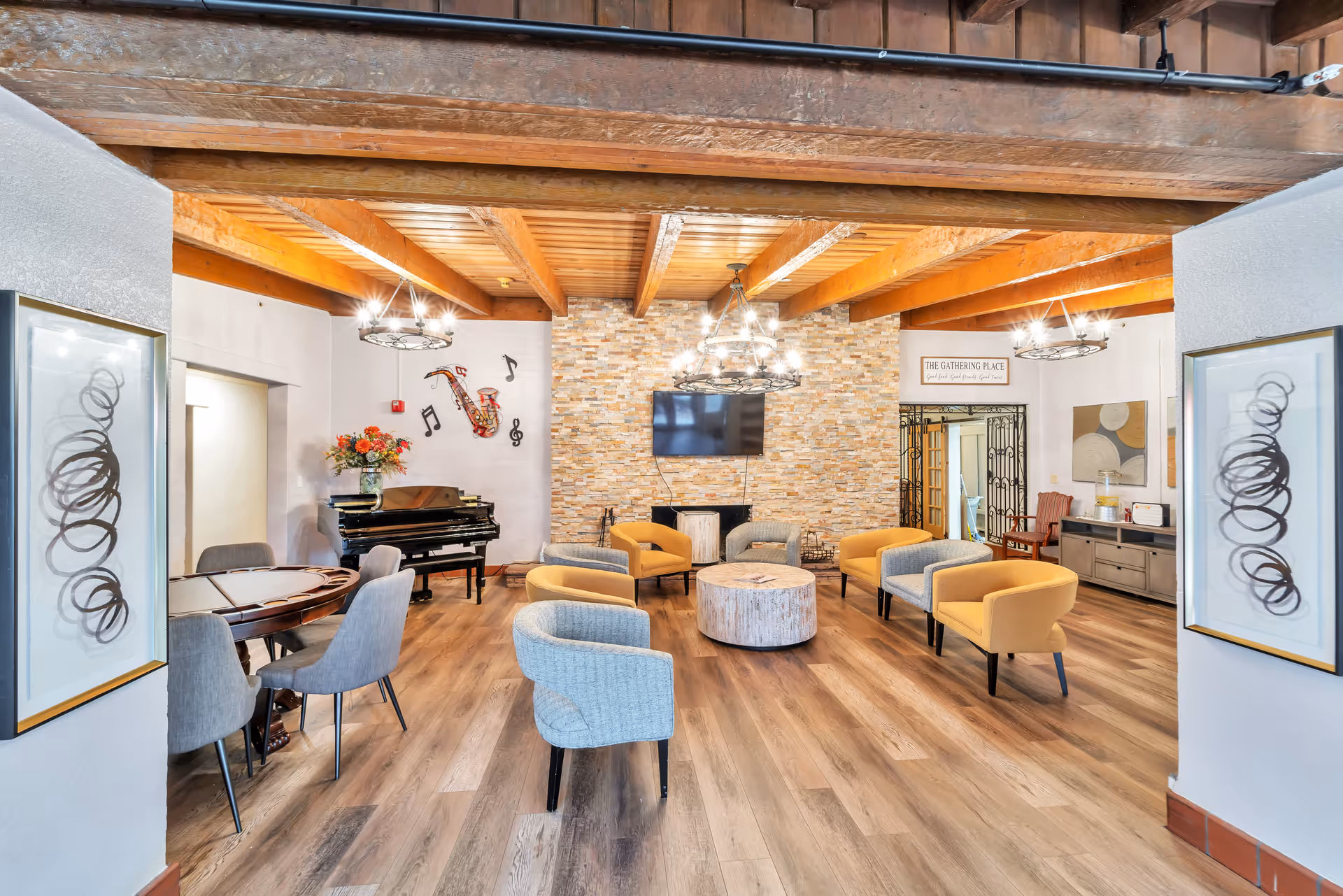 A spacious senior living common area with wooden ceiling beams and a stone accent wall featuring a mounted flat-screen TV. The room is furnished with a mix of yellow and gray armchairs arranged around a round wooden coffee table. On the left side, there is a black piano with a vase of flowers on top and musical note wall decorations. Two modern chandeliers hang from the ceiling, and framed abstract art pieces are displayed on the walls. A sign above a doorway reads 'The Gathering Place'.