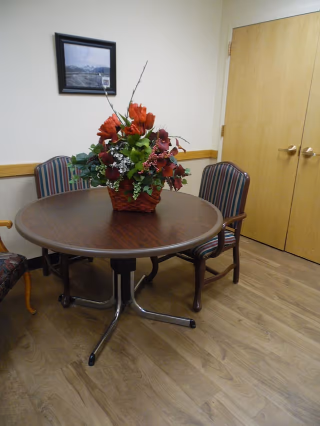 A small round wooden table with a floral arrangement centerpiece featuring red and green flowers in a woven basket. The table is surrounded by three chairs with striped upholstery. The room has light-colored walls, a wooden floor, a framed picture on the wall, and a set of double wooden doors.