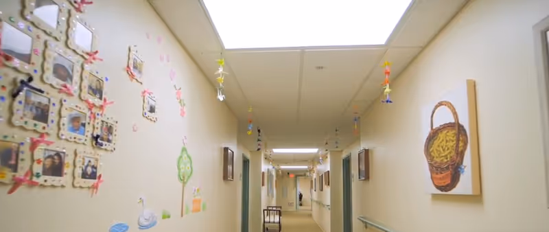 A well-lit hallway in a senior living facility decorated with colorful hanging ornaments from the ceiling and framed photos and artwork on the walls. The floor is carpeted, and there are handrails along the walls for support.