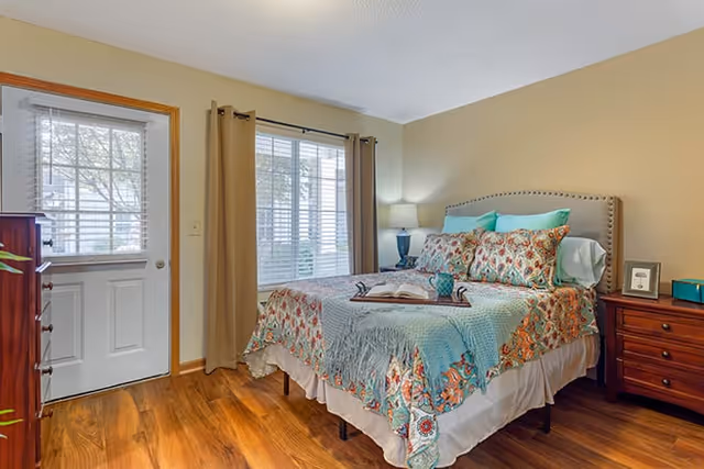 A cozy bedroom with a bed featuring a colorful floral bedspread and multiple pillows. There is a wooden nightstand with a lamp and picture frame on the right side of the bed. A window with blinds and beige curtains is next to a white door with a window, and the floor is wooden.