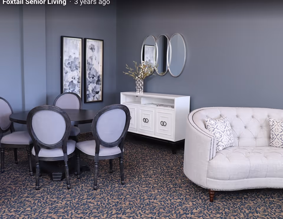 A cozy interior space featuring a round dark wood dining table surrounded by five upholstered chairs with light gray cushions. To the right, there is a curved beige tufted sofa with patterned throw pillows. Against the wall, a white sideboard with three cabinet doors is topped with a decorative vase holding branches. Above the sideboard, three round mirrors are mounted on a gray wall, and two vertical framed abstract artworks hang nearby. The floor is covered with a patterned carpet in dark and light tones.