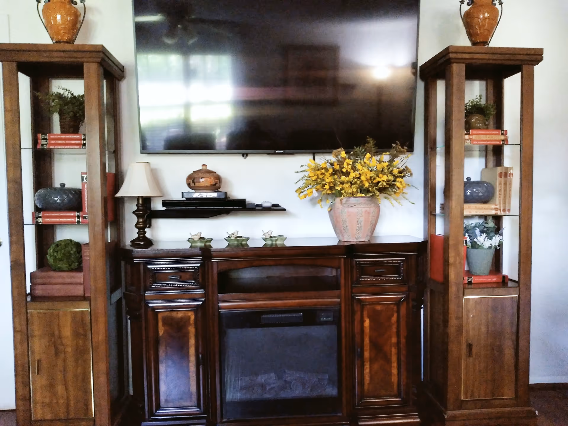 A living room setup featuring a large flat-screen TV mounted on the wall above a dark wooden cabinet with a built-in electric fireplace. On top of the cabinet are decorative items including a table lamp, a small wooden container, three small bird figurines, and a large vase with yellow flowers. Flanking the cabinet are two tall wooden display shelves with glass panels, holding various decorative items such as books, plants, and pottery.