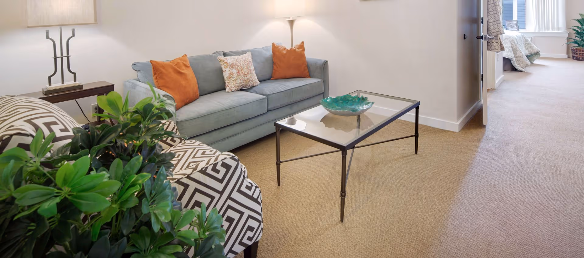 A bright living room with a light-blue sofa, patterned armchair, glass coffee table and plants, opening into a bedroom area.