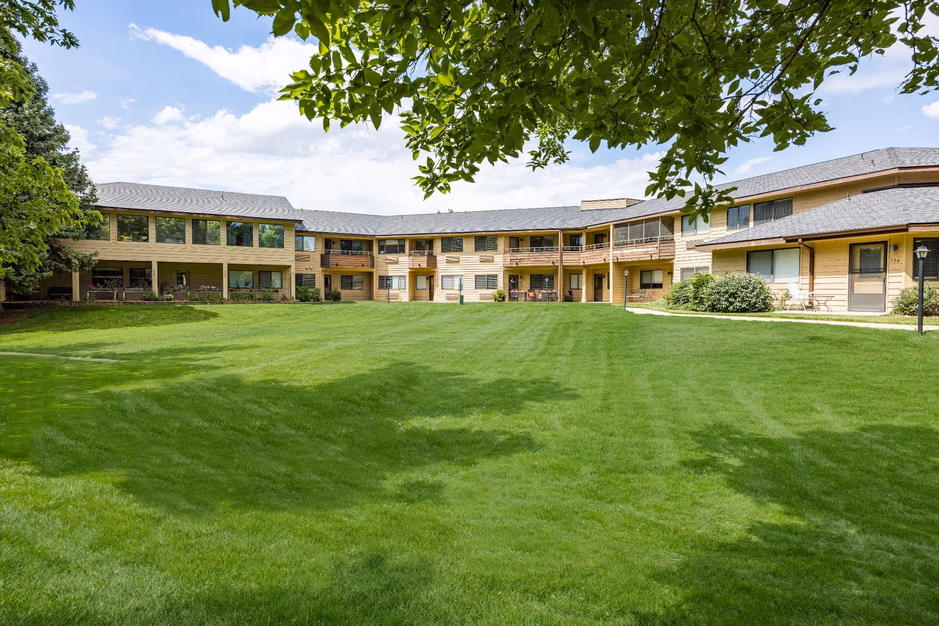 A wide green lawn and courtyard in front of a two-story senior living building with balconies and surrounding trees.