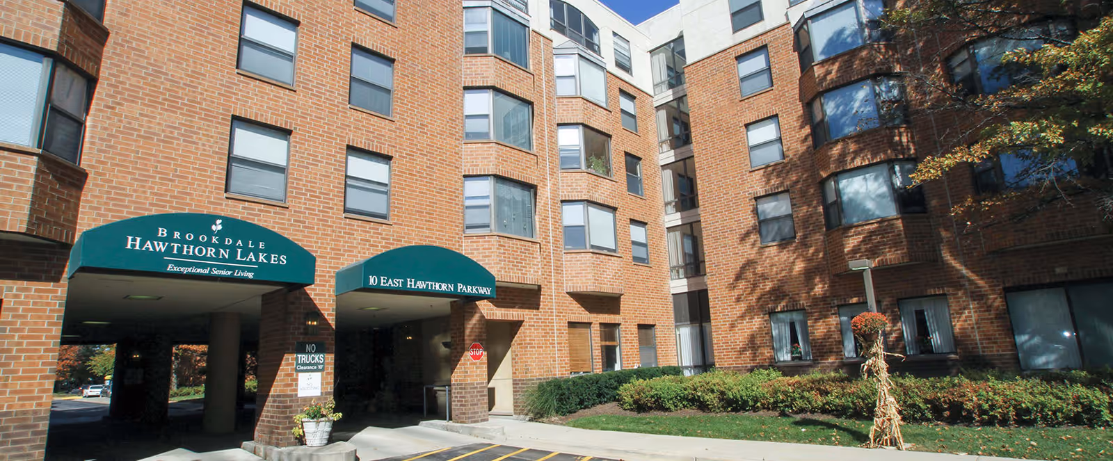 Exterior view of a multi-story brick senior living facility with green awnings over the entrances. The awnings read 'Brookdale Hawthorn Lakes Exceptional Senior Living' and '10 East Hawthorn Parkway'. There are windows on each floor and some landscaping with bushes and a small tree in front of the building.