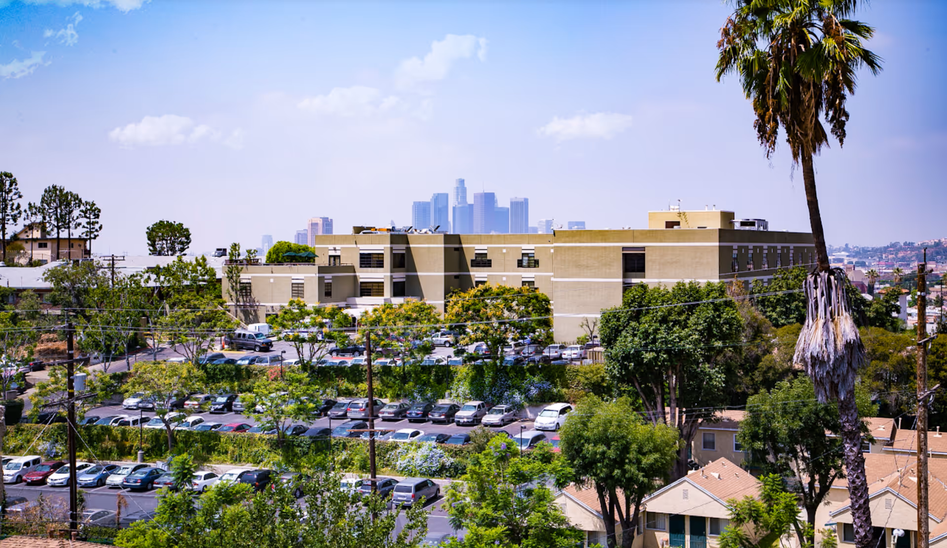View of Kei-Ai Los Angeles Healthcare Center building with multiple floors, surrounded by trees and a parking lot filled with cars. In the background, the skyline of downtown Los Angeles is visible under a partly cloudy sky.