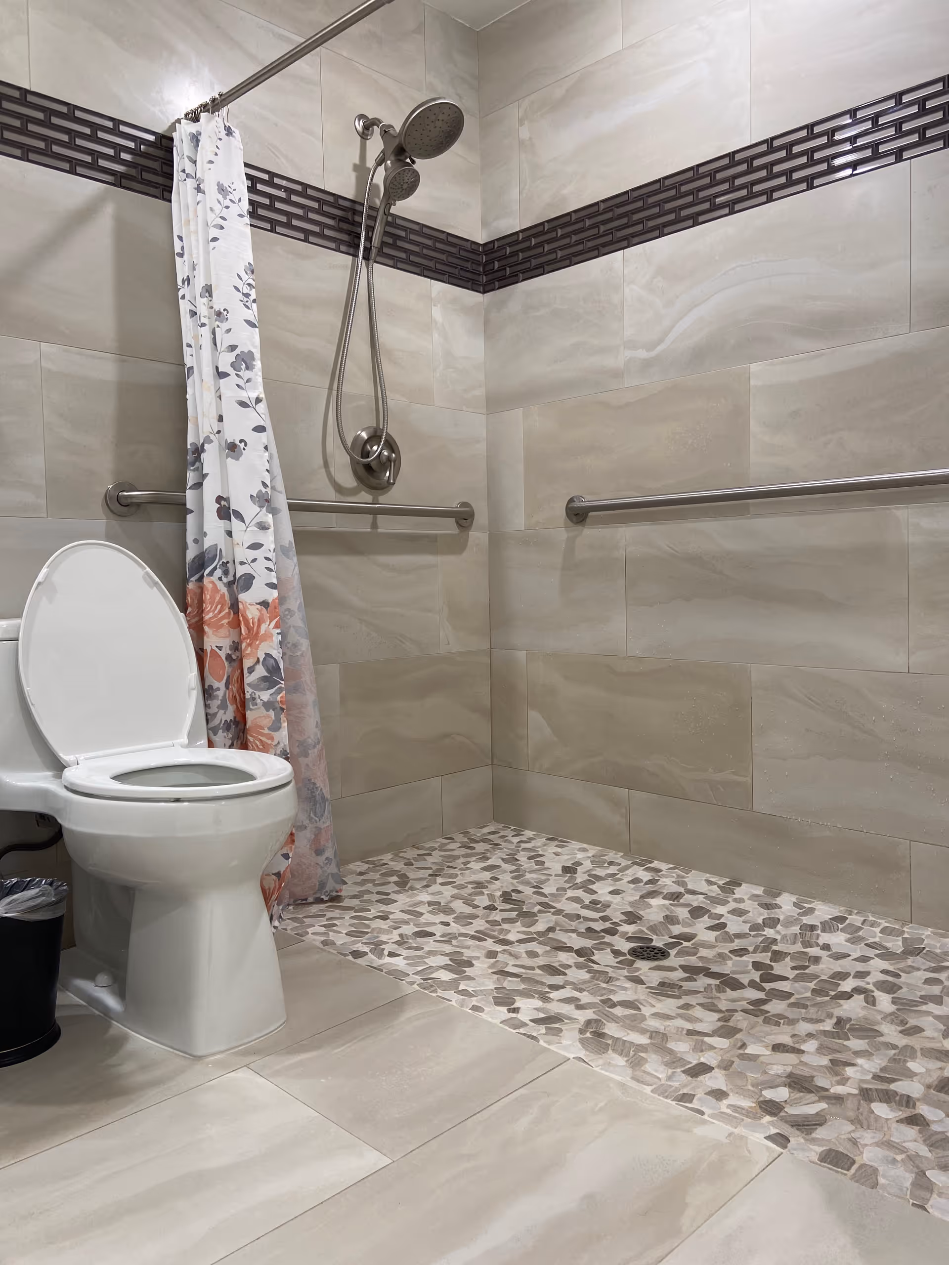 A bathroom with beige tiled walls and floor, featuring a white toilet with the lid open, a floral shower curtain, a walk-in shower area with pebble-style floor tiles, a handheld showerhead, and two horizontal grab bars on the walls.