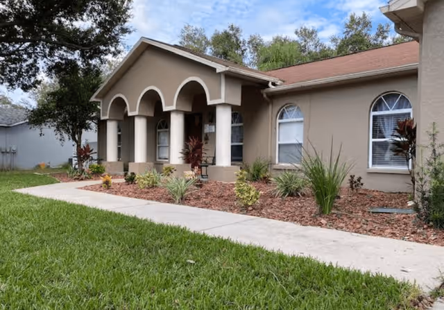 Exterior view of a single-story beige building with a red roof, featuring an arched porch supported by columns. The front yard has a concrete walkway, green grass, and landscaped plants with mulch. Trees are visible in the background under a partly cloudy sky.