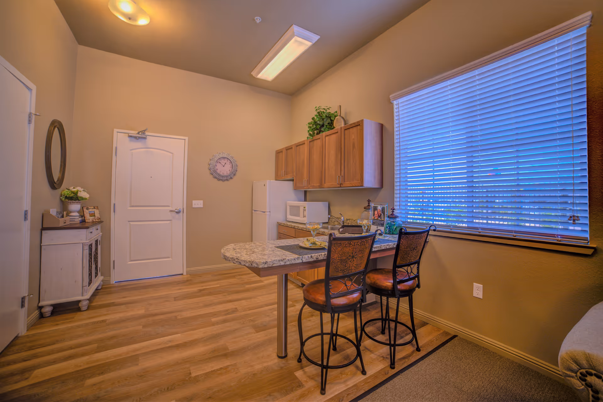 Interior view of a small kitchen area in a senior living facility with wooden cabinets, a white refrigerator, microwave, and a granite countertop island with two high chairs. There is a large window with blinds on the right side and a door on the left wall. The floor is wooden, and there is a small cabinet with decorative items near the door.