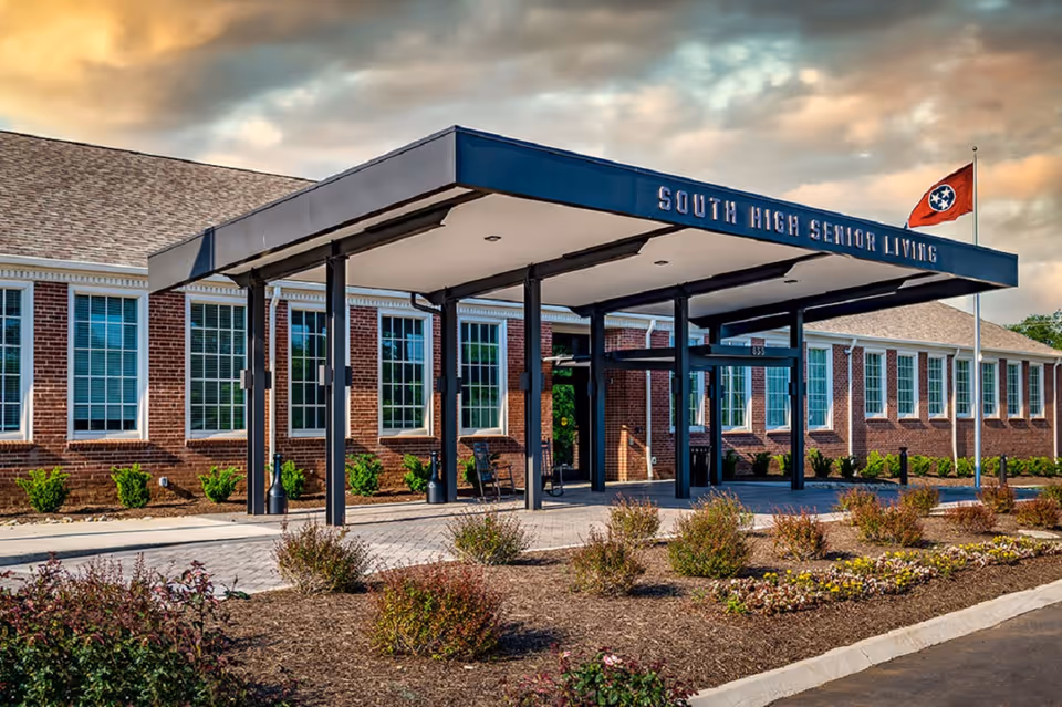 Front entrance canopy of a brick senior living building reading 'South High Senior Living' with landscaping and a flagpole.