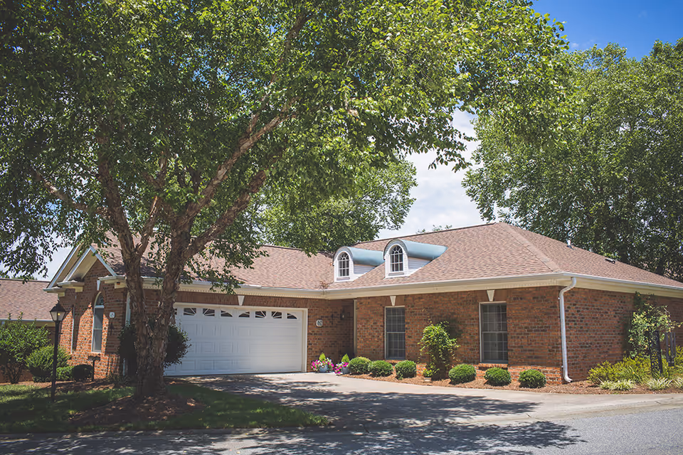 Exterior view of a single-story brick building with a two-car garage, surrounded by green trees and shrubs under a partly cloudy sky.