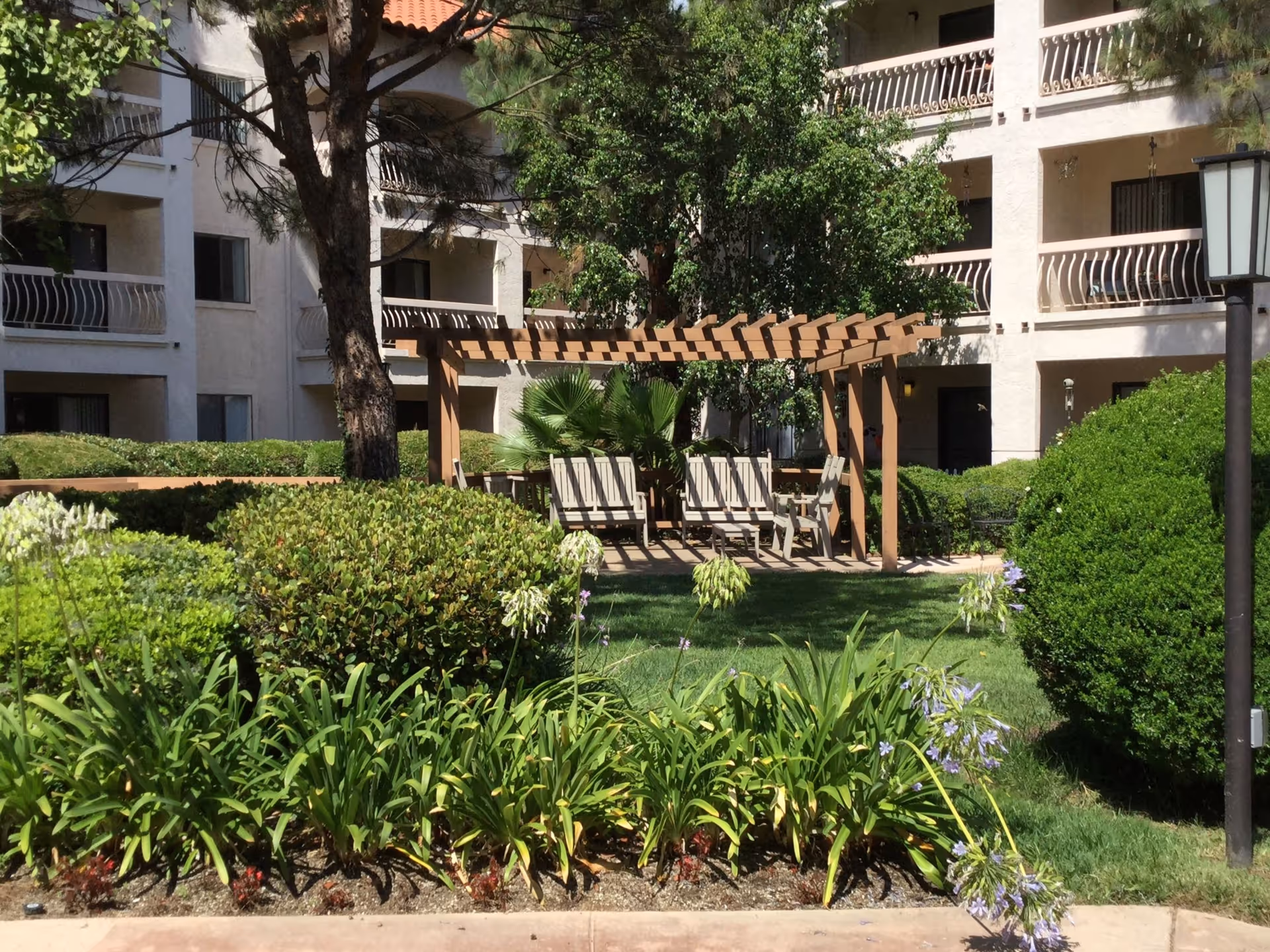 Outdoor garden area at Solstice Senior Living at El Cajon featuring a wooden pergola with several chairs underneath, surrounded by green bushes, trees, and flowering plants. The background shows a multi-story building with balconies.