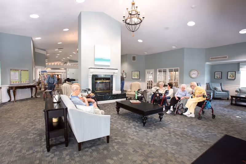 A spacious senior living facility common area with several elderly residents seated on sofas and chairs around a central black coffee table. The room features a fireplace with a painting above it, soft blue walls, and a chandelier hanging from the ceiling. Some residents use walkers and are engaged in conversation. The area is well-lit with recessed lighting and has a carpeted floor.