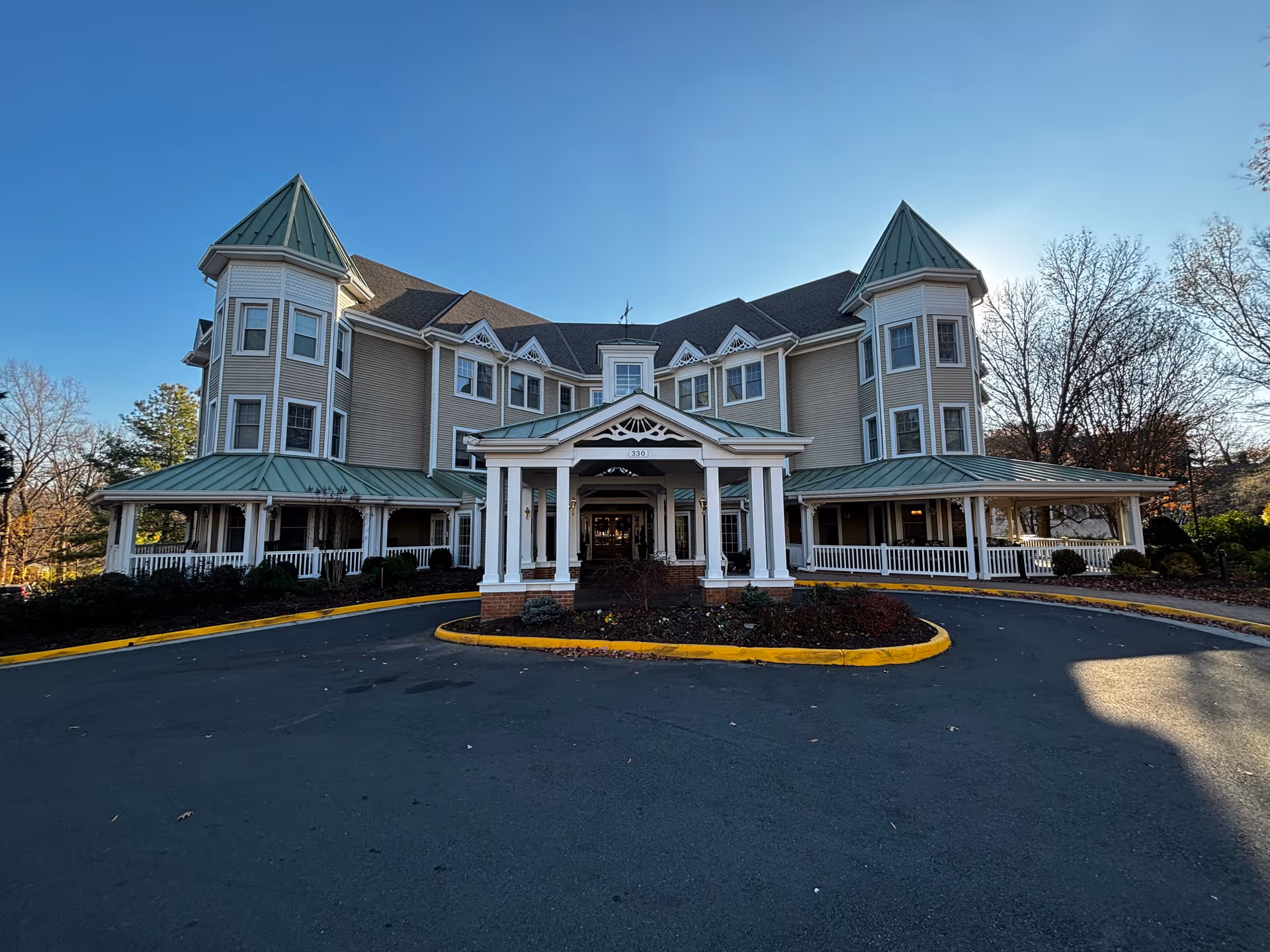 Front view of a large three-story senior living building with green metal roofs, wraparound porches, and a covered entrance driveway.