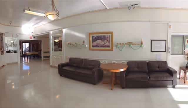 A well-lit interior common area with two dark brown leather sofas separated by a round wooden table. The walls are white with decorative molding and a framed picture hanging above the sofas. There are green garlands hanging on the wall and a chandelier light fixture on the ceiling. In the background, there is a hallway leading to another room with more seating visible.