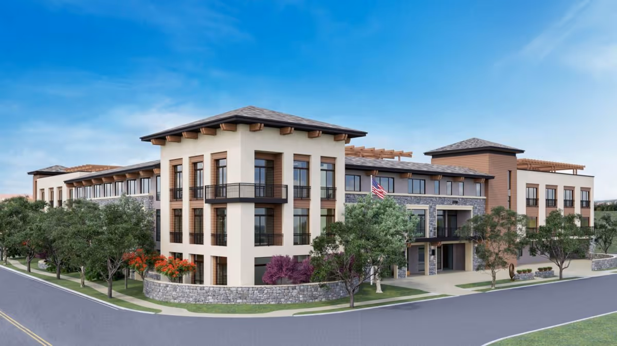Exterior view of a modern three-story senior living facility building with large windows, stone and stucco facade, surrounded by trees and landscaping, under a clear blue sky.