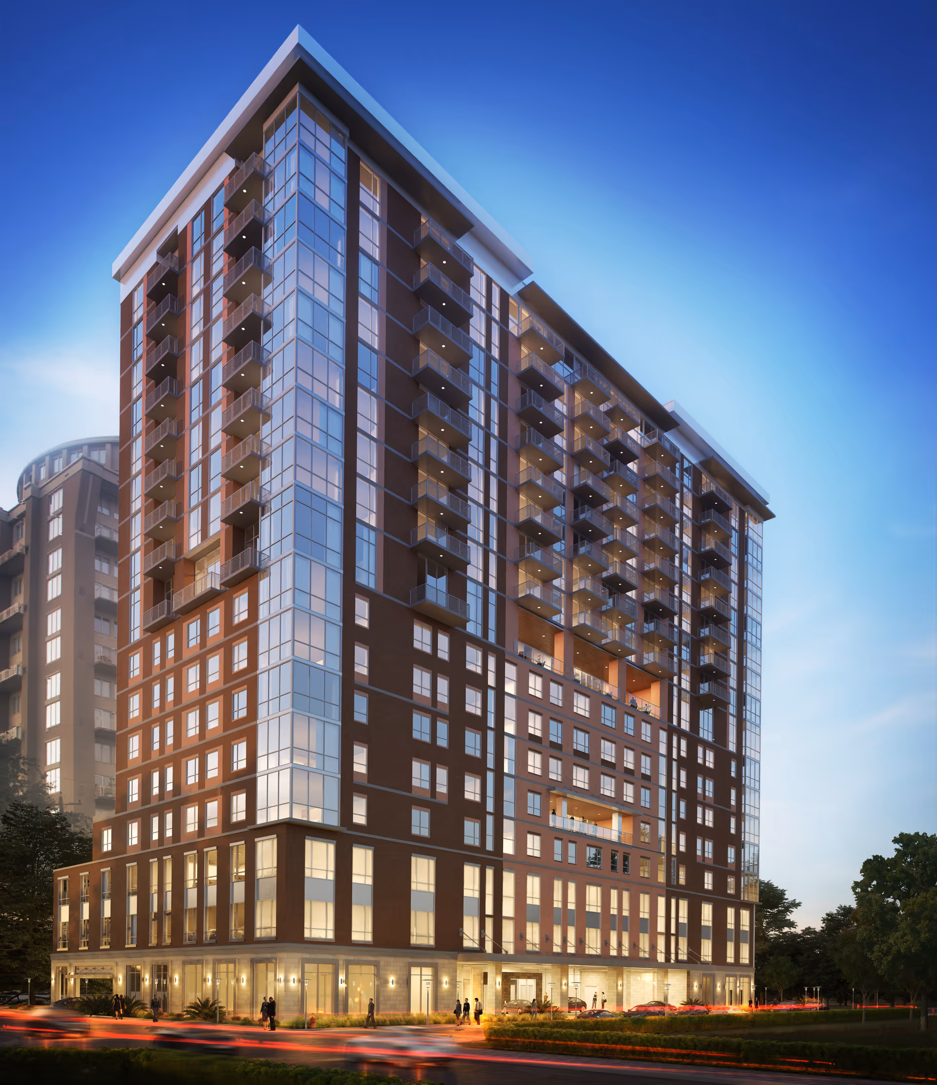 A modern multi-story residential building with large glass windows and balconies, illuminated at dusk with people walking near the entrance and cars passing by on the street.