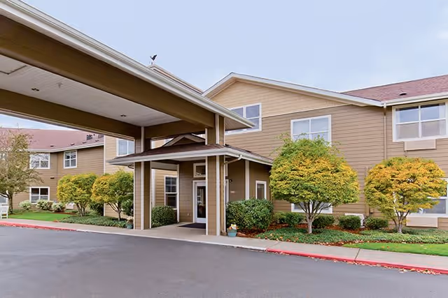 Exterior view of Heron Pointe Senior Living facility showing a two-story building with beige siding, multiple windows, a covered entrance with a driveway, and landscaped greenery including trimmed bushes and small trees.