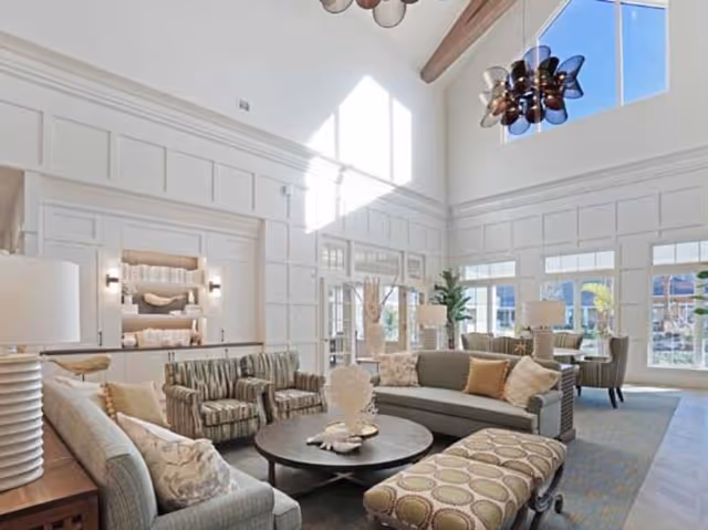 A spacious and bright living room with high ceilings and large windows letting in natural light. The room is furnished with multiple sofas, armchairs, a round coffee table, and an upholstered bench. Decorative elements include table lamps, cushions, and plants, creating a comfortable and inviting atmosphere.
