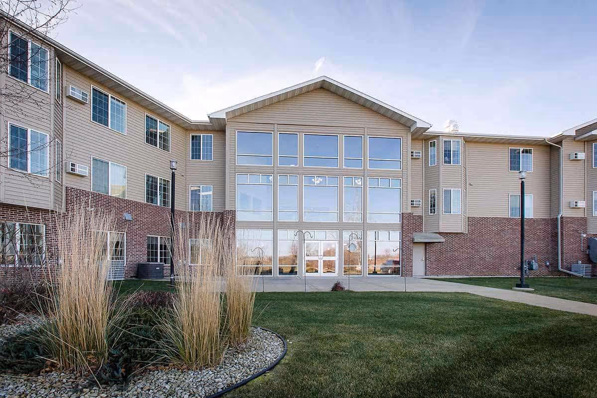 Exterior view of a three-story senior living facility building with large windows and a landscaped lawn with ornamental grasses and shrubs in front.