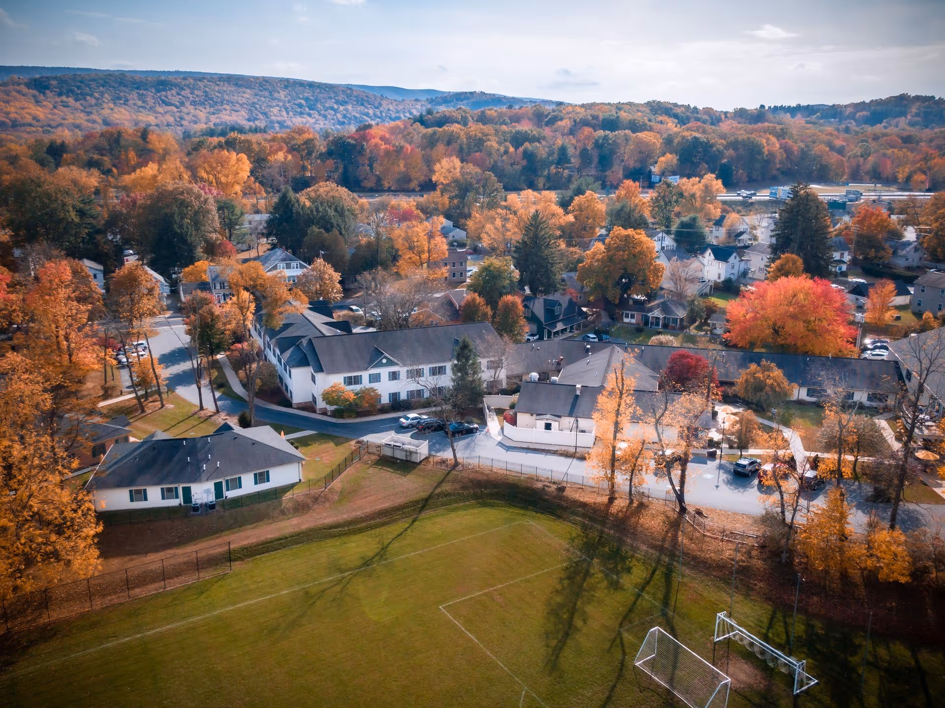 Aerial view of a senior living facility named Grace Park Senior Living surrounded by autumn trees with colorful fall foliage, residential houses, and a grassy sports field with soccer goals in the foreground.
