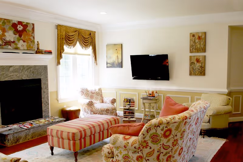 A cozy living room with floral and striped upholstered furniture including a sofa, armchairs, and an ottoman. There is a fireplace with a marble surround on the left, a window with gold curtains, a wall-mounted flat-screen TV, and framed artwork on the walls. The room has warm lighting and a soft area rug on the floor.