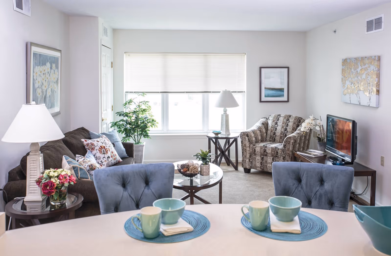 A bright and cozy living room with a brown sofa adorned with patterned cushions, a patterned armchair, a flat-screen TV on a wooden stand, and a large window with blinds letting in natural light. There are two side tables with lamps and decorative plants, a round glass coffee table with small plants and decor, and a dining counter in the foreground set with blue placemats, bowls, and mugs.