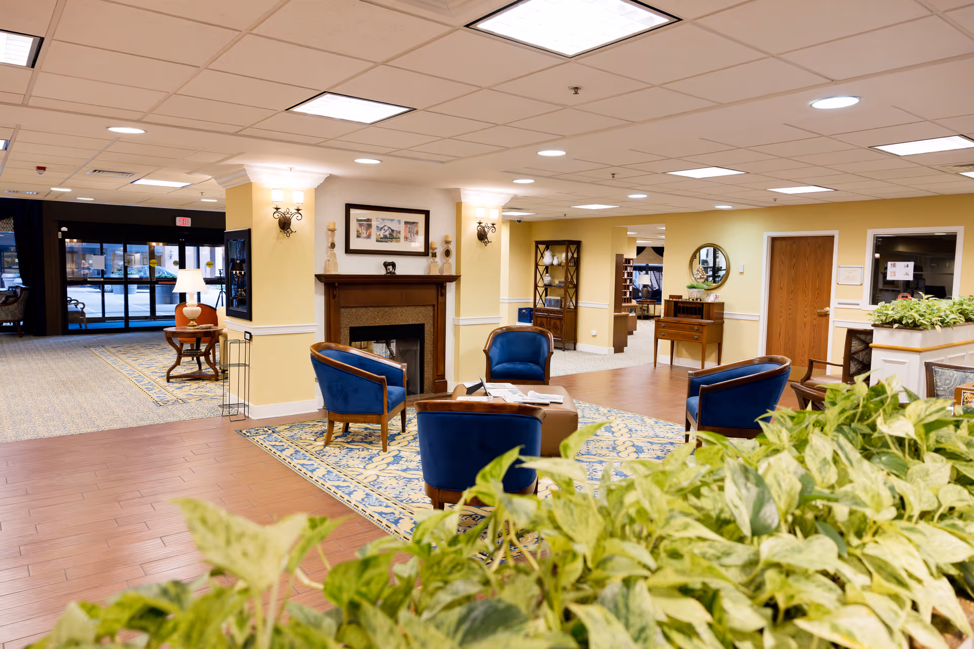 A bright and welcoming senior living facility common area with a seating arrangement of four blue upholstered chairs around a small table on a patterned rug. There is a fireplace with decorative items and framed artwork above it. The room has yellow walls, wooden flooring, and ceiling lights. In the foreground, there are green plants, and in the background, there is an entrance with glass doors and additional seating and furniture.