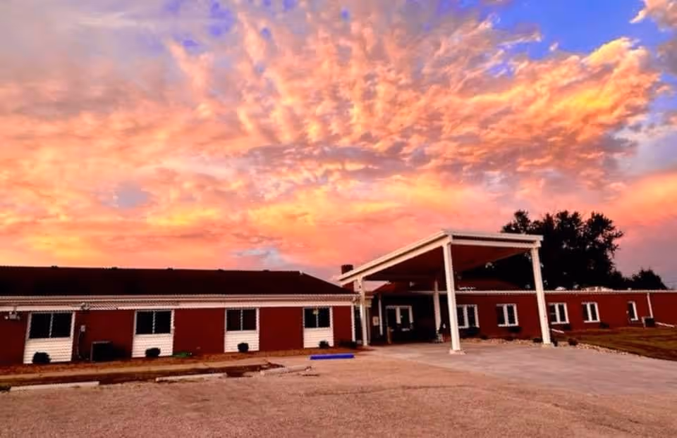 Single-story red brick building with a covered entrance beneath a colorful sunset sky.