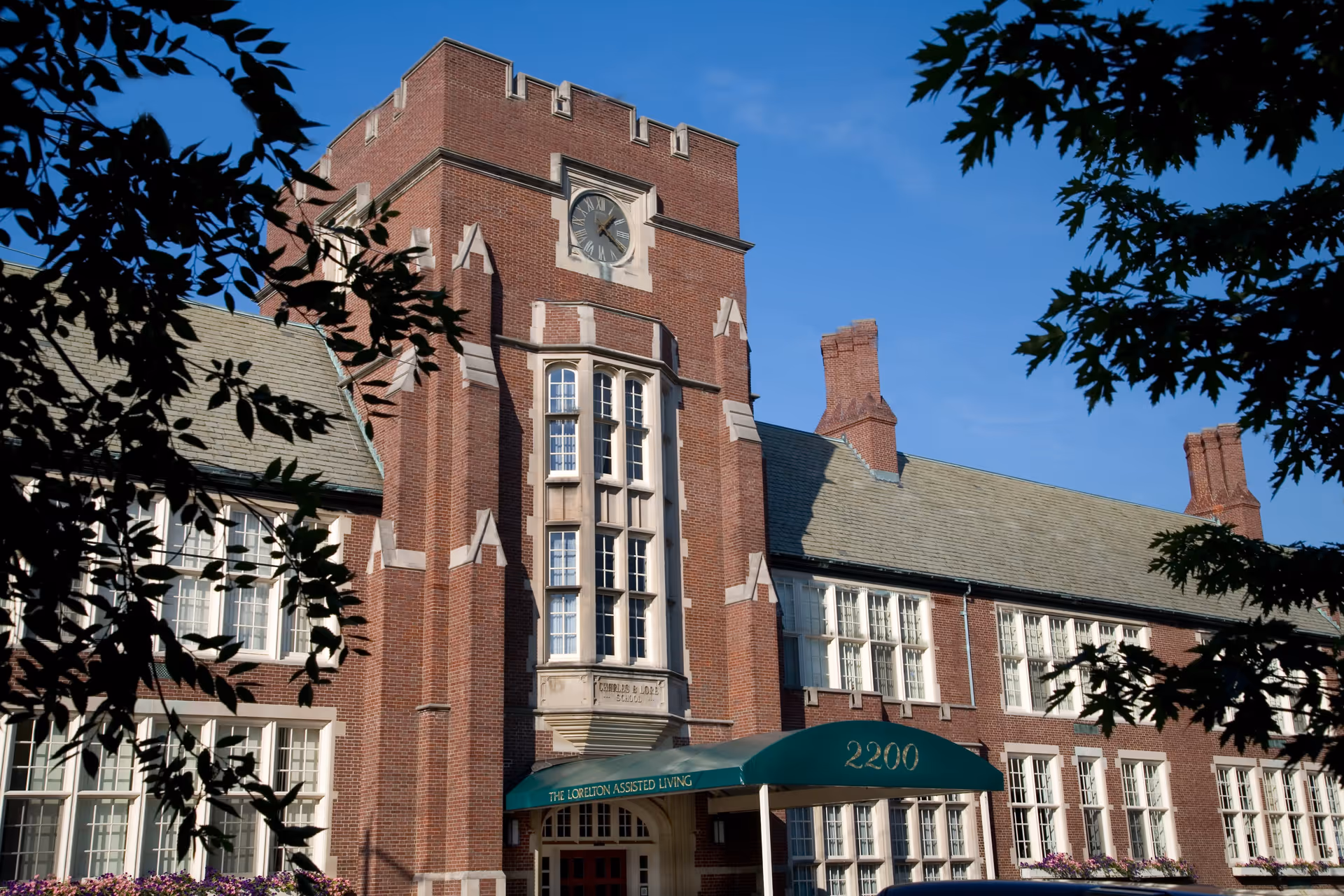 Exterior view of The Lorelton building, a large brick structure with a clock tower, multiple windows, and a green awning over the entrance displaying the address 2200 and the words 'The Lorelton Assisted Living'. The building is framed by tree branches and set against a clear blue sky.