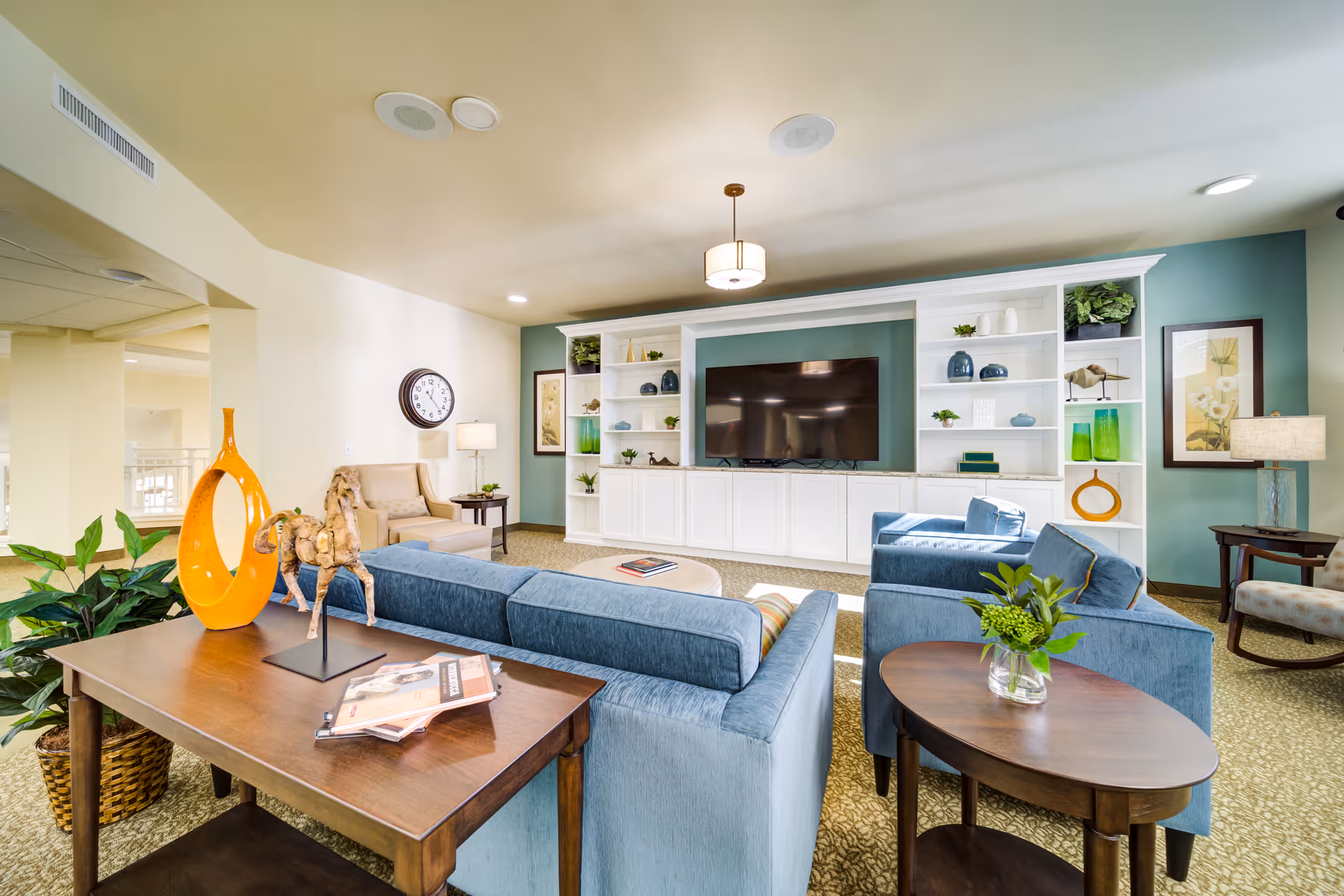 A bright and spacious living room with light blue sofas and armchairs arranged around a central coffee table. The room features a large built-in white shelving unit with decorative items and a flat-screen TV mounted in the center. There are two side tables with lamps, framed artwork on the walls, and a wooden console table with decorative sculptures and magazines. The carpet is patterned, and the walls are painted in soft neutral and teal tones.