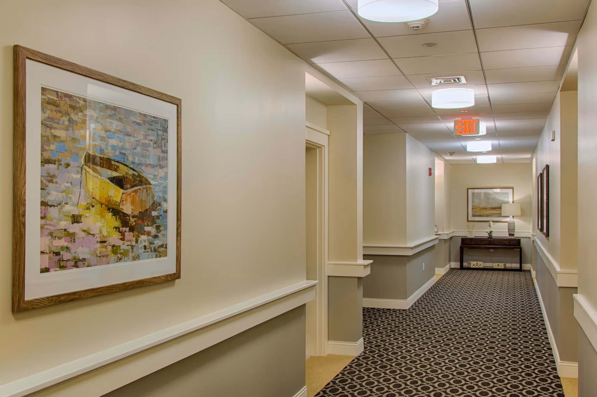 A well-lit hallway in a senior living facility with patterned carpet, beige and gray walls, framed artwork on the walls, and a small table with a lamp and decorative items at the end of the corridor.