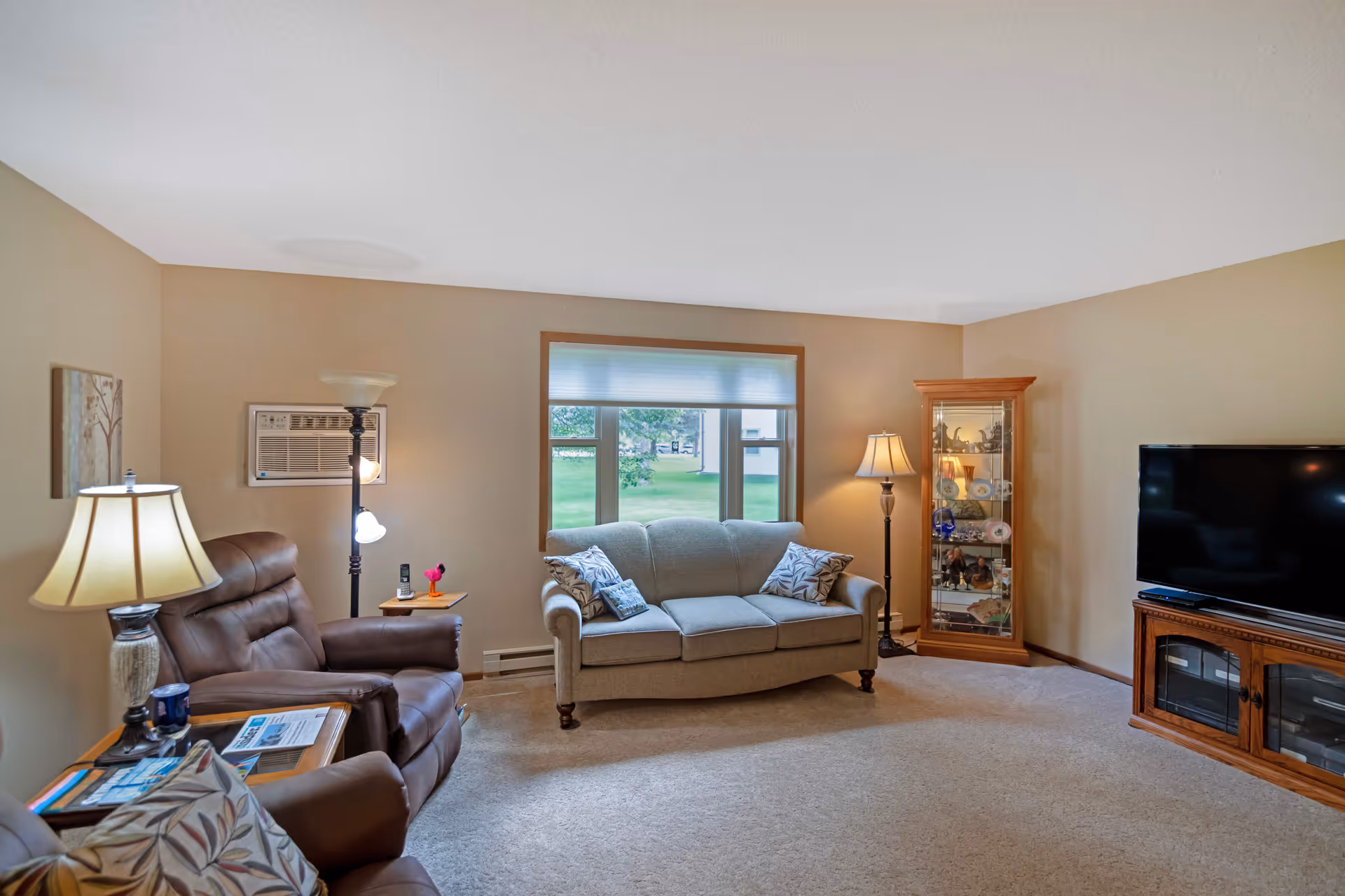 Comfortable living room with a sofa, two recliner chairs, a TV on a wooden stand, floor lamps, and a window overlooking a green lawn.