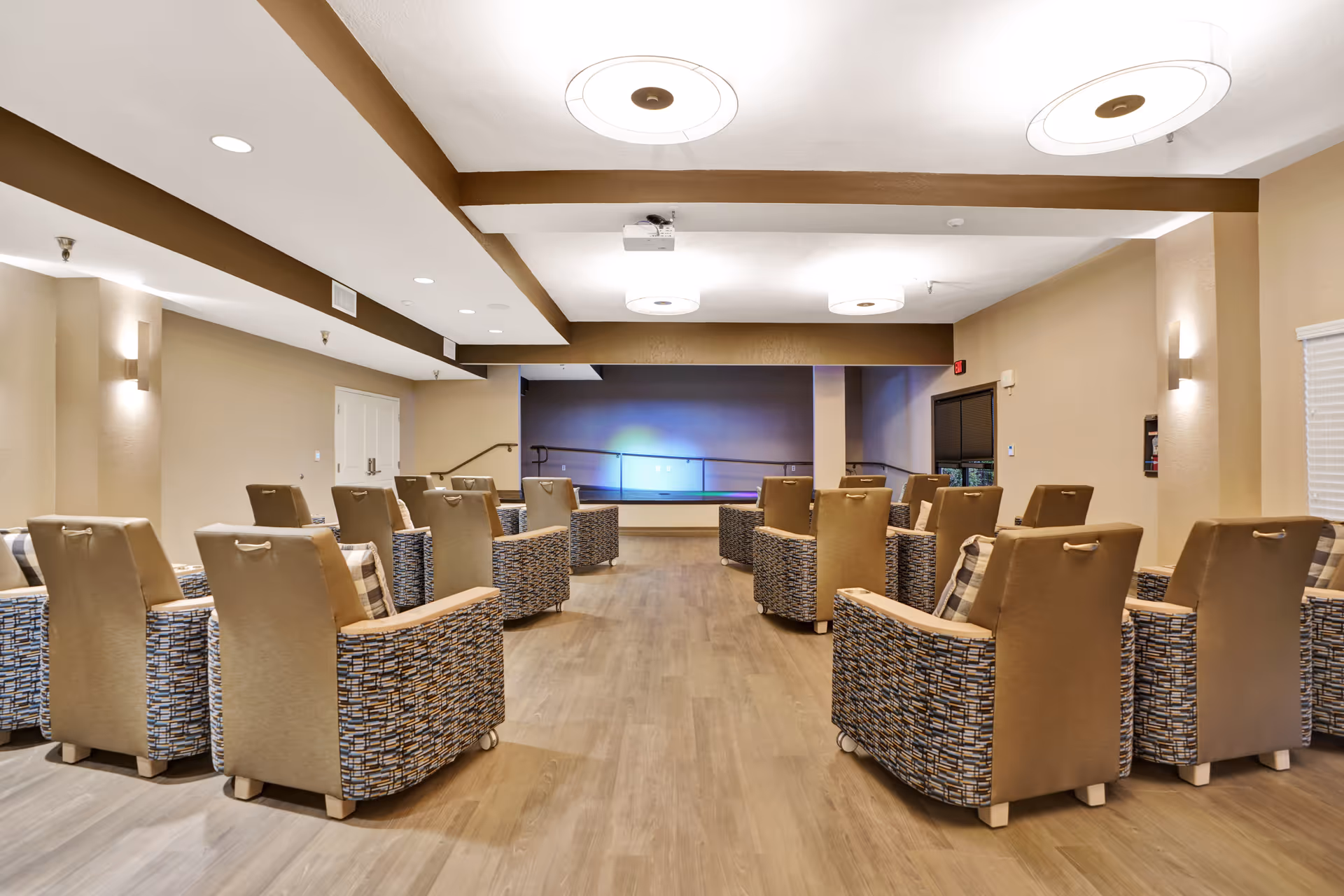 A spacious room with multiple cushioned armchairs arranged in rows facing a small stage area with a ramp. The room has wooden flooring, beige walls, and modern ceiling lights. The setting appears to be a common area or lounge in a senior living facility.