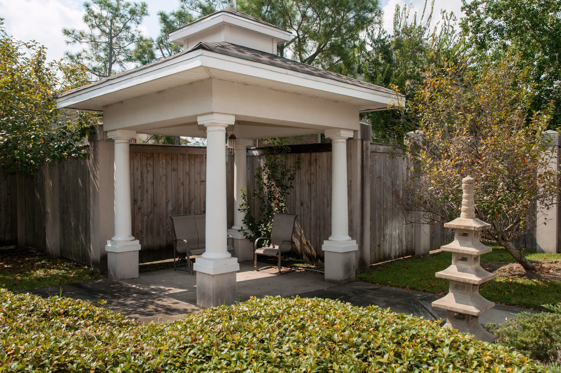 A small pavilion with white columns and seating in a garden courtyard beside a wooden fence and a decorative stone pagoda.