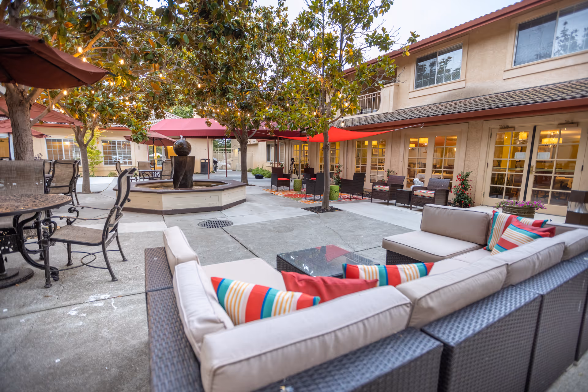 Outdoor courtyard area at Cogir of North Bay Senior Living featuring cushioned wicker seating with colorful pillows, metal tables and chairs, large red umbrellas, trees, and a central water fountain. The building with large windows and doors surrounds the courtyard.