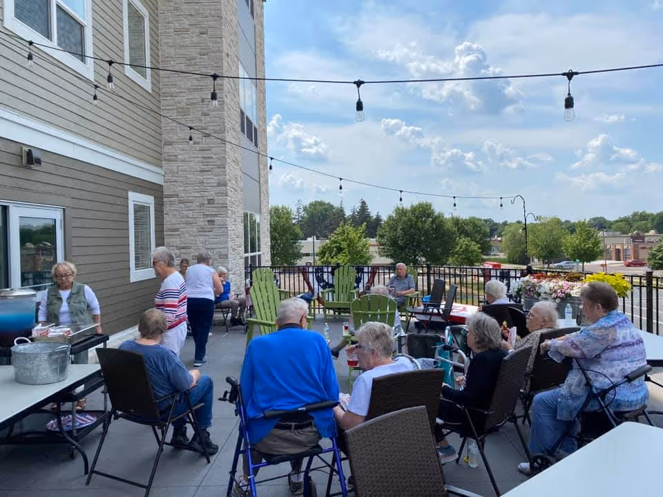 A group of elderly people sitting and socializing on an outdoor patio at a senior living facility. The patio has string lights overhead, green Adirondack chairs, and a table with refreshments. Trees and buildings are visible in the background under a partly cloudy sky.