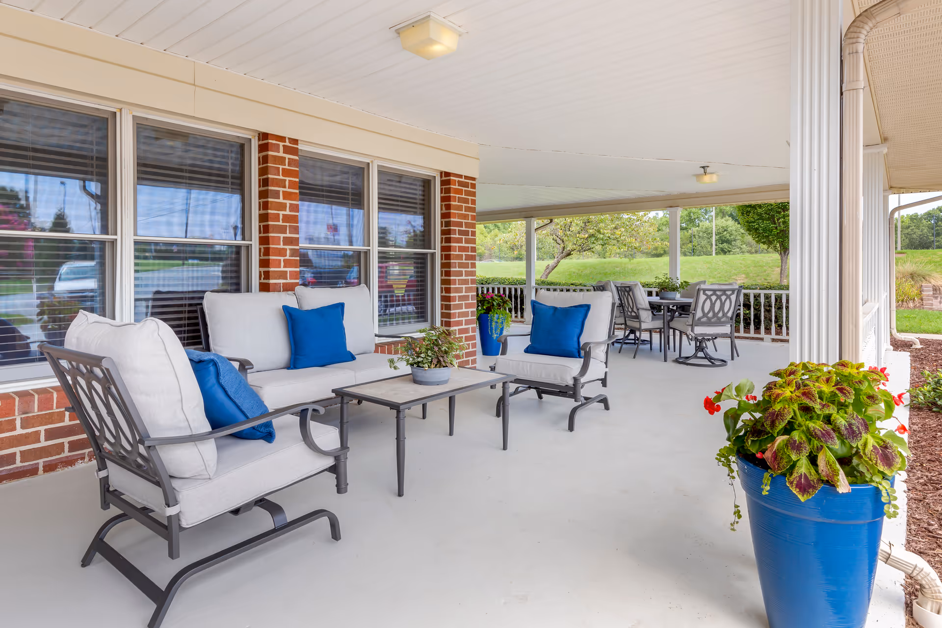 Covered porch with cushioned metal patio seating and blue pillows, a coffee table, dining set, and potted plants overlooking a lawn.