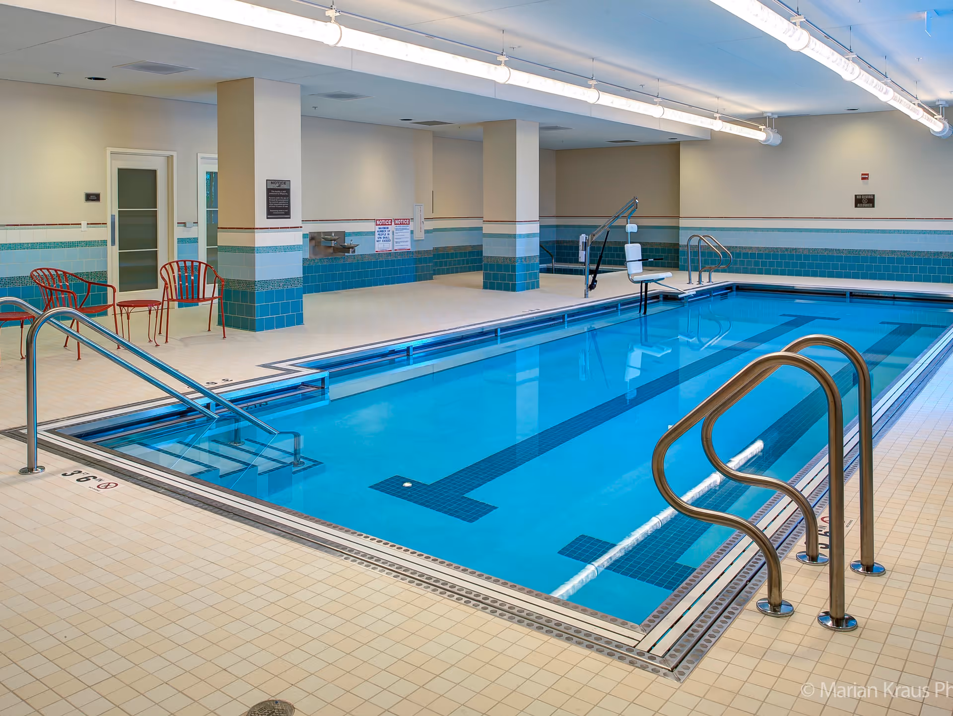 Indoor swimming pool with clear blue water, metal handrails, pool lift chair, and red chairs along the tiled poolside area. The walls are decorated with blue and white tiles, and there are overhead fluorescent lights.