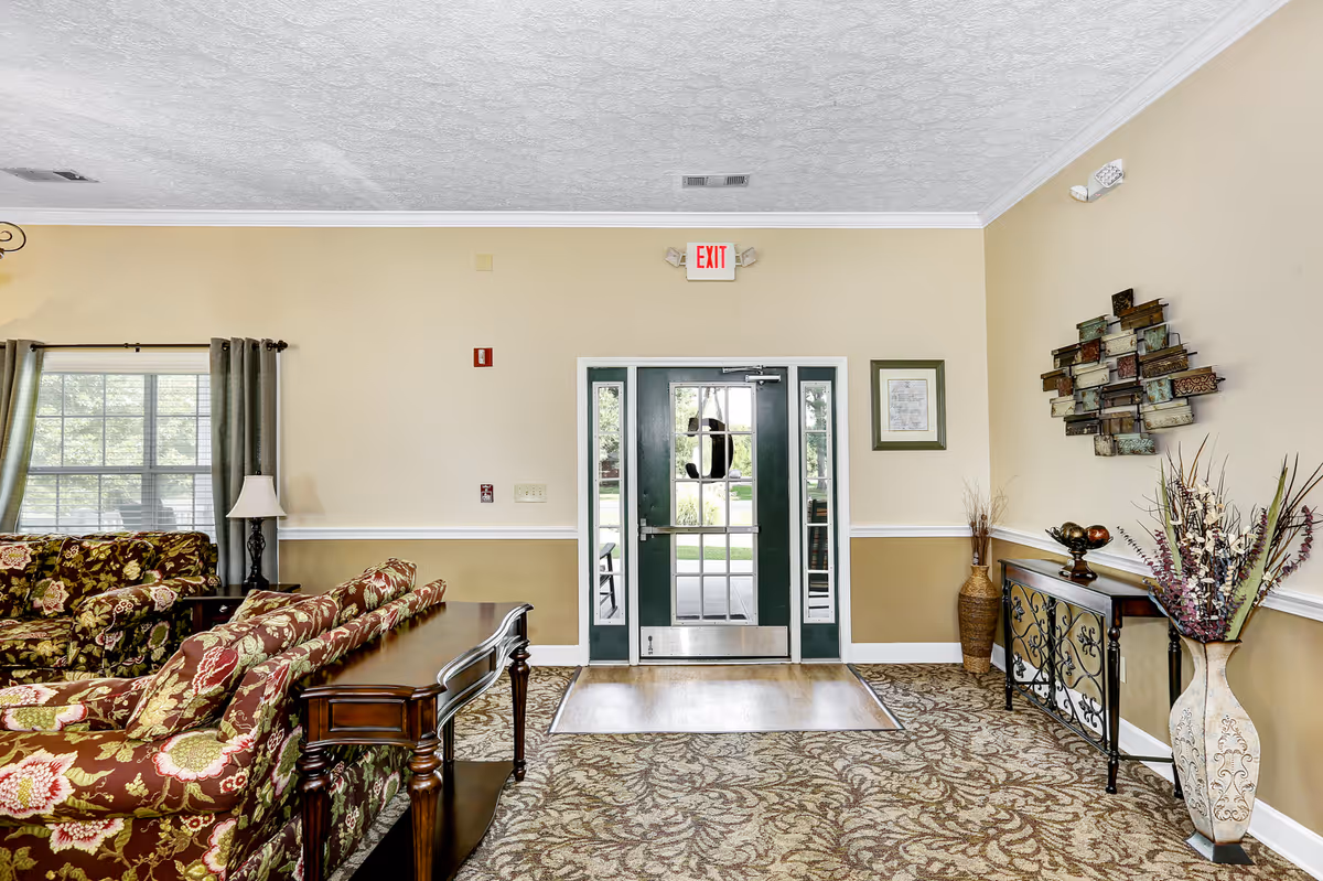Interior view of a living room area in a senior living facility with floral patterned sofas, a wooden console table, decorative vases with plants, a wall art piece, and a glass door exit leading outside.