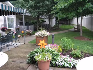 Outdoor garden area with a paved walkway, green grass, trees, and various potted and planted flowers. There is a metal table and chairs set under a striped awning attached to a building on the left side. A bench is visible along the path in the background.