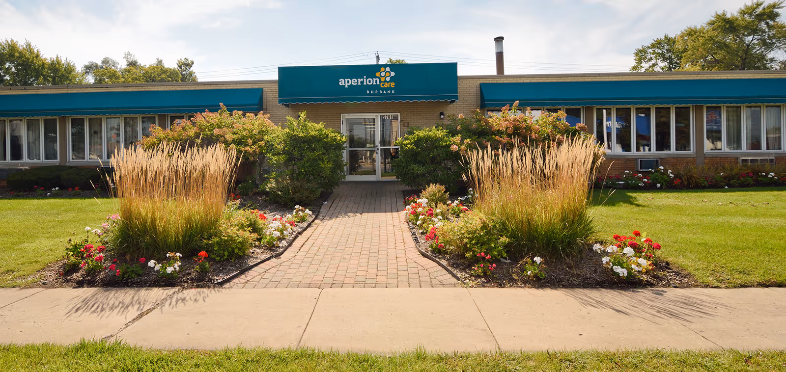 Entrance of a single-story brick senior care building with a green awning, central glass doors, and landscaped flowerbeds along a brick walkway.