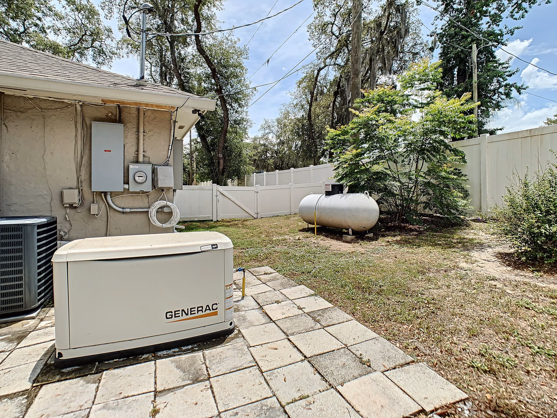 Outdoor area of a facility showing a Generac generator on a paved surface next to a building wall with electrical panels and wiring. There is a large propane tank on the grass near a white fence, with trees and bushes in the background under a partly cloudy sky.