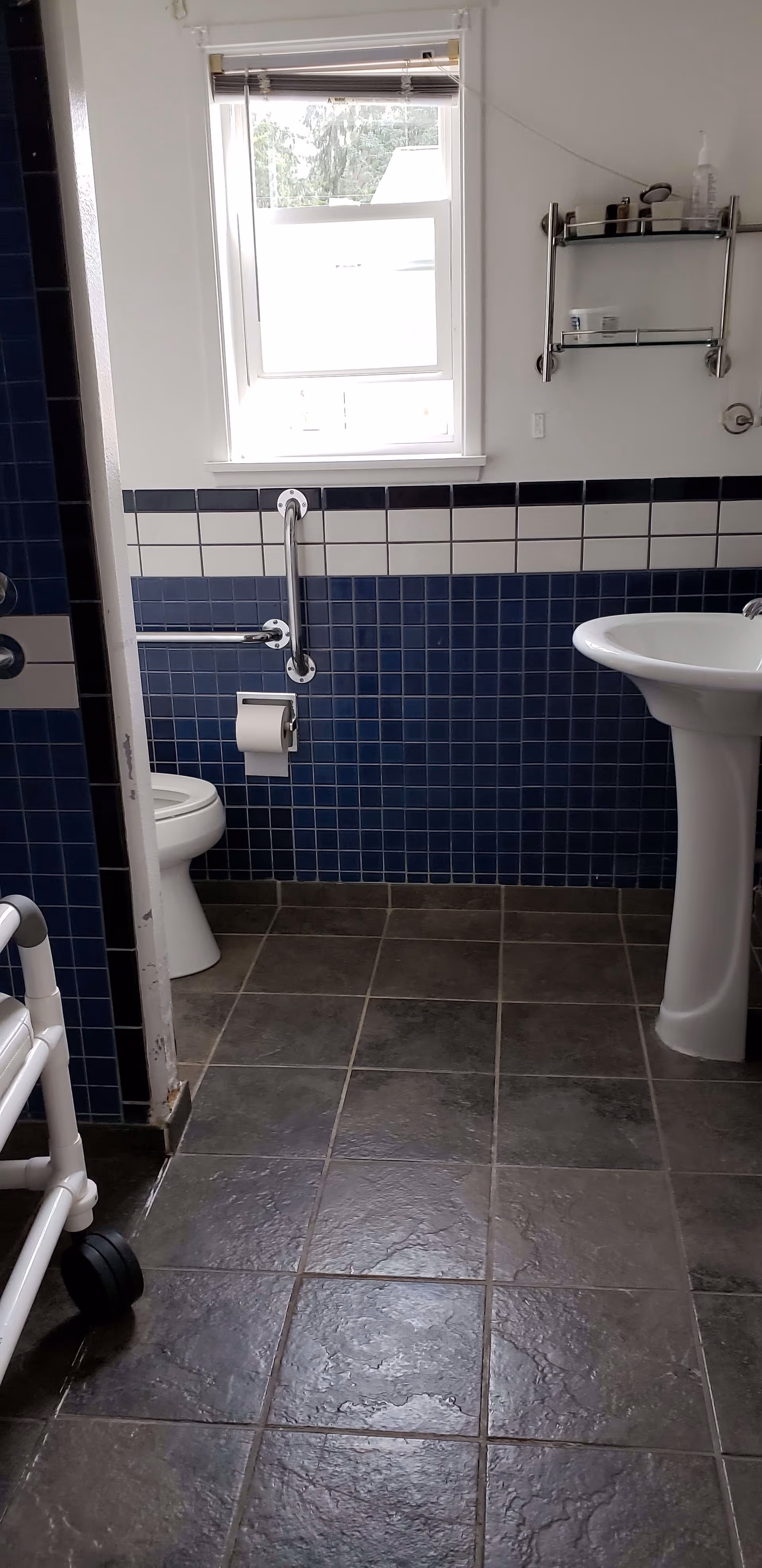Bathroom with dark blue and white tiled walls, a white toilet with a grab bar and toilet paper holder, a white pedestal sink, a window with blinds, a metal shelf with toiletries, and a walker partially visible on the left side.